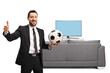 © Ljupco Smokovski - Man in a black suit showing thumbs up and holding a soccer ball in front of tv and sofa
