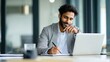 © Johannes - young indian businessman entrepreneur using computer working online doing research taking notes watching business training webinar in office having virtual chat meeting looking at laptop at work no l