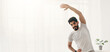 © Prostock-studio - Covid-19 pandemic, workout with gadget at home and healthy lifestyle. Attractive young bearded arabian man doing warm-up or stretching for arms in minimalist living room interior, copy space