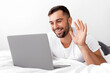 © Prostock-studio - Happy relaxed millennial caucasian muscular guy lies on white bed, communicates and waves his hand at computer in bedroom interior. Video call, meeting and rest at home in free time during covid-19