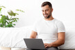 © Prostock-studio - Cheerful attractive caucasian millennial male to home in white t-shirt works on laptop sits beside bed in white bedroom interior. Freelance, home business and independent contractor due covid-19