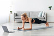 © Prostock-studio - Online training with coach remotely, new normal and social distance during covid-19. Happy millennial female in sportswear doing plank on mat on floor in minimalist living room interior with laptop