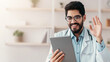 © Prostock-studio - Modern video conference or online consultation with doctor in clinic. Happy young arabian bearded guy in white coat and glasses holds tablet, waves hand and talk, sits at table in office interior