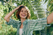 © SHOTPRIME STUDIO - Joyful young woman with a wide smile stands under tropical palm fronds, wearing a green striped shirt, capturing a bright, carefree moment.