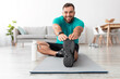 © Prostock-studio - Cheerful young handsome guy fitness blogger with beard doing stretching or warm-up for legs on mat with bottle of water in living room interior. Workout at home, health care, sport, covid-19 outbreak