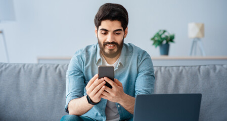 Business and telecommuting concept. Happy young arab man using smartphone and modern laptop computer, reading text message, sitting at desk with gadgets, working at home, copy space
