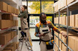 © DC Studio - Black employee checking cardboard boxes with goods on shelving units, working with inventory list in a small scale depot. Ensuring small business in house operations, electronic services.