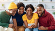 © Prostock-studio - Happy african american fellows sitting on couch at home, using mobile phone together, laughing, having video conference with friends. Positive black men and women watching funny videos on smartphone