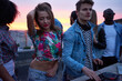 © pressmaster - Young Caucasian woman dancing with hand in hair beside man operating DJ equipment at rooftop party with diverse group of young adults during sunset