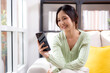 © NT_studio - Young asian businesswoman looking smartphone smiling and relax sitting on sofa in living room at home, cheerful woman watching smart phone sitting on couch in home, lifestyle and communication.