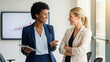 © rawrsiu - Two businesswomen, one African American and one Caucasian, are having a conversation in a modern office setting with a graph displayed on a screen in the background.