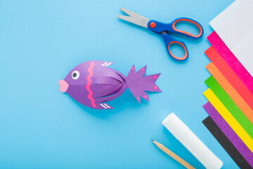 Wall Mural - Purple fish shape, scissors, glue stick, pencil and colorful application paper on light blue table background. Pastel color. Closeup. Child handcraft. Top down view.