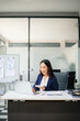 © laddawan - Confident Asian businesswoman writing on clipboard, smiling while working at desk with laptop and documents.