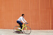 © pressmaster - Young adult Latin man riding bicycle along orange corrugated wall, wearing sunglasses and business attire, commuting outdoors in urban setting, hands gripping handlebars, focused expression