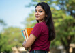 © Brijesh - Confident Latin Indian Female Student in Maroon T-Shirt Holding Books Outdoors | College Campus Portrait with Blue Sky and Greenery