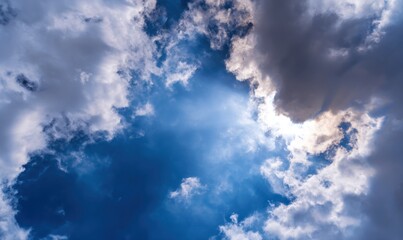 Wall Mural - Dramatic upward view of a sunny blue sky with fluffy white, grey and black clouds, sunlight streaming down