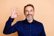 © deagreez - Confident senior businessman in blue shirt smiles and gestures with hand on beige background, expressing positivity