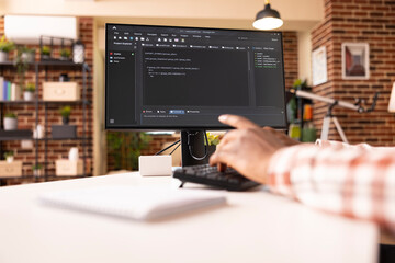 Closeup of computer screen showing programming scripts in progress in brick wall apartment. African American web developer seated at desk, writing code on desktop pc in brick wall home.