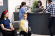 © DC Studio - Caucasian little girl sits calmly in hospital waiting area while asian man completes payment at front desk. Modern clinic lobby showing mother and child waiting for medical care.