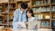 © bongkarn - Glasses asian woman student sitting and holding tablet showing to a man friend at library's table