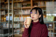 © bongkarn - Glasses asian woman holding or drinking coffee and looking away while sitting at table in library