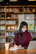 © bongkarn - Smiling glasses asian woman writing in notebook as looking at laptop aside coffee at library's table