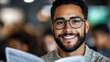© Yuliia - Young mixed race professional man with glasses and beard wearing grey sweater smiling confidently at camera against blurred office background.