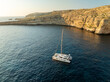 © AmazingAerialAgency - Aerial view of a lone yacht sailing in the deep blue waters near the rugged Dwejra cliffs, where the sun kisses the horizon, Dwejra cliffs, Gozo, Malta.