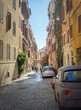 Narrow cobblestone street in Rome is lined with traditional buildings. The walls are adorned with shutters and climbing plants