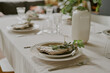 © AnnaStills - Closeup showing elegant table setting with stacked plates, linen napkins, cutlery, drinking glasses and decorative greenery, neutral tablecloth covering dining table surface