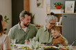 © AnnaStills - Caucasian senior woman embracing smiling child while sitting at dining table with young Caucasian man, family members gathering and sharing meal together indoors