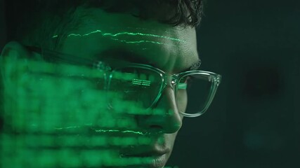 Focused young programmer with green computer code reflecting in his glasses in a dark room
