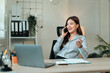 © Crystal - Young Asian businesswoman talking on phone and working on laptop in modern office, smiling and gesturing while discussing business matters