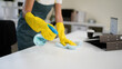© Stella 16 - Young asian woman wearing yellow gloves and an apron, smiling while cleaning a white table with disinfectant spray and a blue cloth, promoting hygiene and cleanliness in her workspace