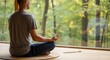 © Feliks - A person sits in a cross-legged pose on a woven mat meditating with hands in a mudra An incense stick burns by a large window overlooking a lush forest