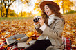 © maxbelchenko - Woman is sitting on blanket in park covered with autumn leaves with camera in hands. Beautiful woman in hat is enjoying autumn landscape and taking pictures on camera. Concept of beauty, relaxation.