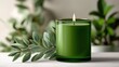 © Tina - A green candle in a glass holder sits next to a sprig of leaves on a light surface. The scene conveys a sense of calm and tranquility.