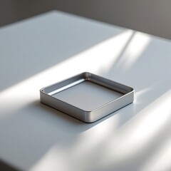 Photo of a minimalist square-shaped mobile holder in matte silver finish placed on a pristine clean white surface with subtle soft reflections and a professional studio glow.