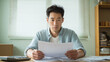 © bephoto - 男性が自宅の机で書類を見て悩むの様子