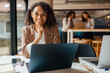© Yaroslav Astakhov - A cheerful professional woman works at her laptop in a stylish office with colleagues in the background.