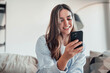 © Daniel - Smiling woman looking at smartphone screen, sitting on couch, enjoying leisure time with mobile device at home, happy young female chatting in social network or surfing internet, shopping online