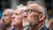 © Muslima - Elderly gentleman with glasses and beard, detailed wrinkles, cinematic close-up, realistic portrait, background crowd softly blurred.