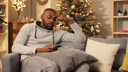 Tired guy in sportive hoodie and jeans sitting on couch on background of decorated Christmas tree with balls and toys. Attractive young man browsing and chatting. Indoors.