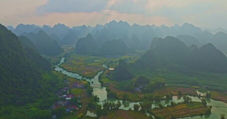 Wall Mural - Aerial view of a riverside village at sunset, with golden light reflecting on flowing waters and distant mountains rising beyond the clouds.Rural Hamlet Surrounded by Mountains and Flowing River