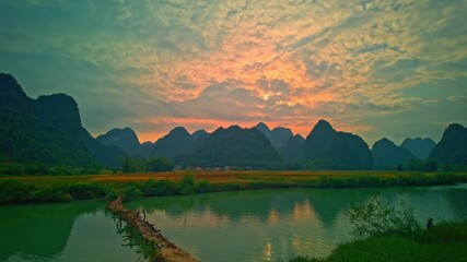 Wall Mural - The calm stream reflects the fiery colors of the setting sun as a wooden bridge connects the fields, with lush greenery and majestic peaks completing this peaceful countryside scene.
