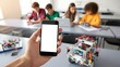 © Jessika Cahya - A person holds a smartphone with a blank white screen in the foreground while students in the background engage in a robotics and coding class.