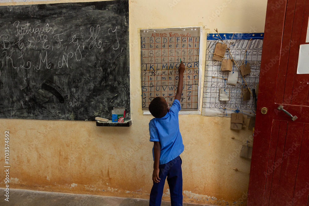 Sparse Classroom A young African boy in a blue shirt points to an alphabet chart on a ...