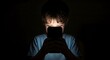 © Qii Stock - Young boy illuminated by the glow of a smartphone screen in a dark room, focusing intently on the device.