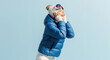 © Vasiliy - A happy young woman in winter ski attire smiles joyfully. Cheerful person in a blue puffer jacket and goggles on a blue background with copy space