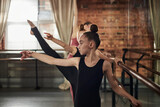 Caucasian girl practicing ballet at barre in dance studio, focusing on arm position while other girls stretching in background, demonstrating concentration and technique during class